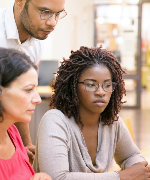 Adult female students consulting instructor in computer class. Man and women sitting and standing at desk, using desktop, looking at monitor, talking. Training concept