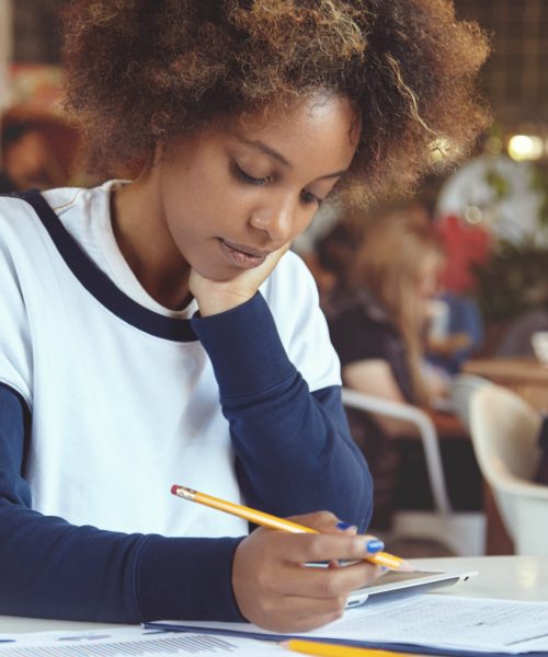 Close-up portrait of tender African female freelancer at cafeteria working on her tablet, writing out something on sheet of paper, looking tired and bored, resting cheek on hand, drinking white coffee